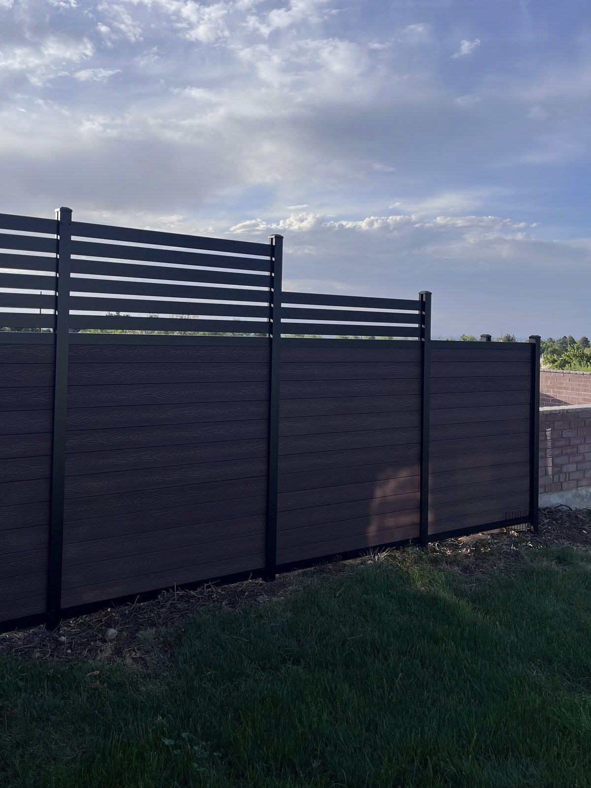 Dark brown horizontal composite fence with black aluminum posts and slatted toppers, stepped in sections, on a Trusted Fence job in the Salt Lake Valley