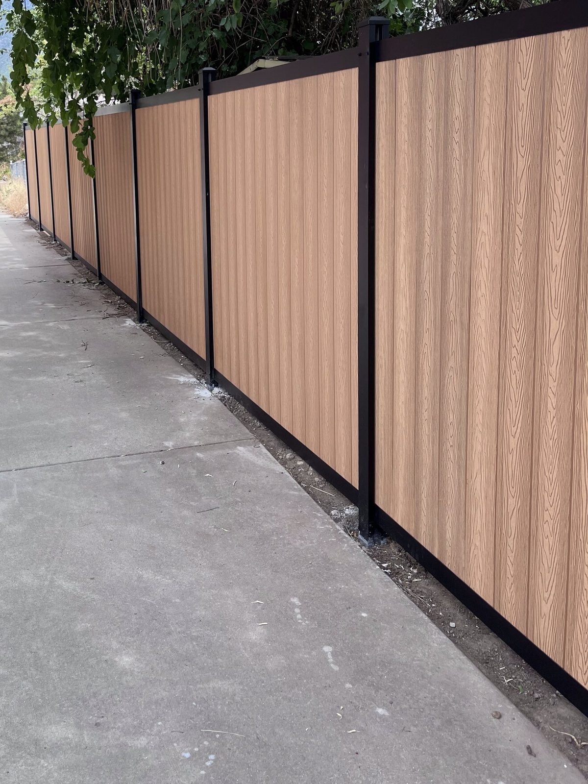 Light brown vertical composite privacy fence with dark aluminum posts along a concrete driveway, on a Trusted Fence job in the Salt Lake Valley