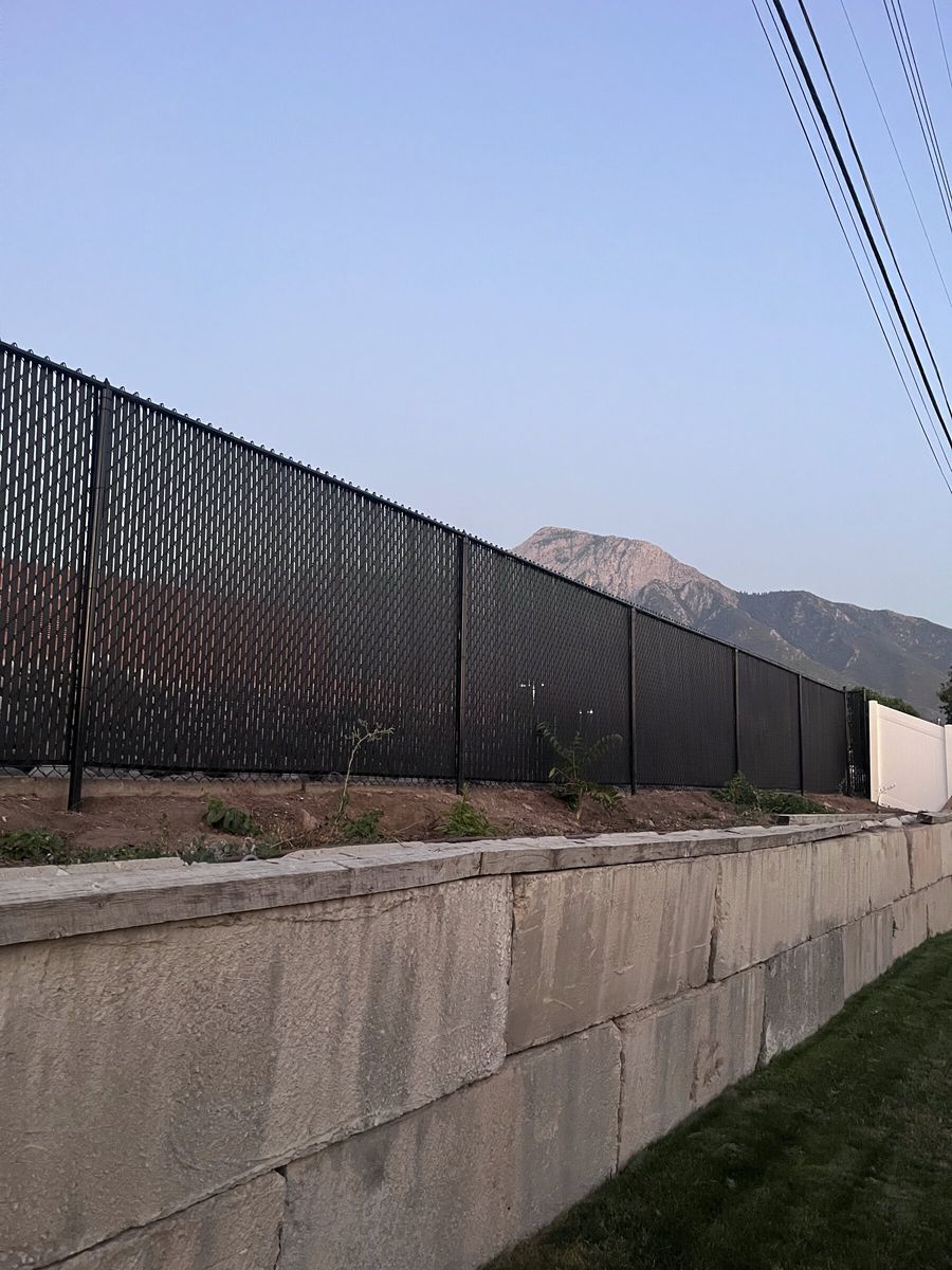 Black chain link fence with privacy slats above a block retaining wall with mountains behind in the Salt Lake Valley