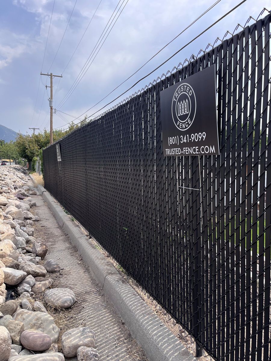 Black chain link fence with privacy slats and a Trusted Fence sign along a landscaped rock edge in the Salt Lake Valley
