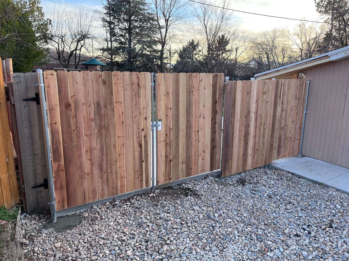 Wood privacy fence with a double drive gate and black hinge across a gravel driveway in Bluffdale, UT
